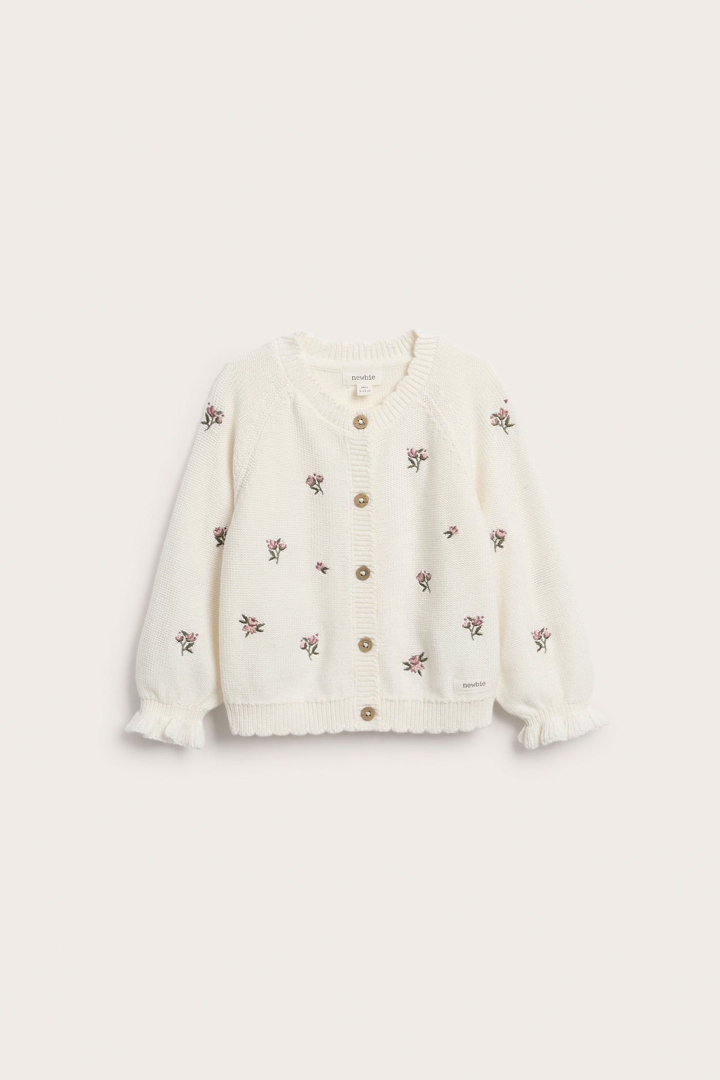 Front view of an off-white knitted cotton kid's cardigan with embroidered pink flowers, button front, scalloped hem, and ruffled cuffs.