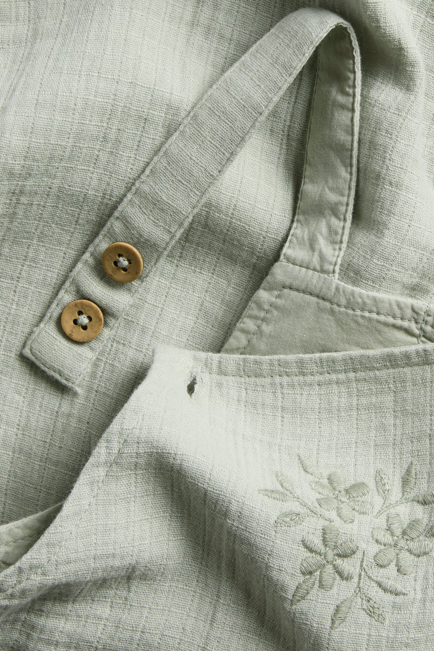 Close-up of a light green cotton baby overall with adjustable wooden button straps and floral embroidery on the chest.