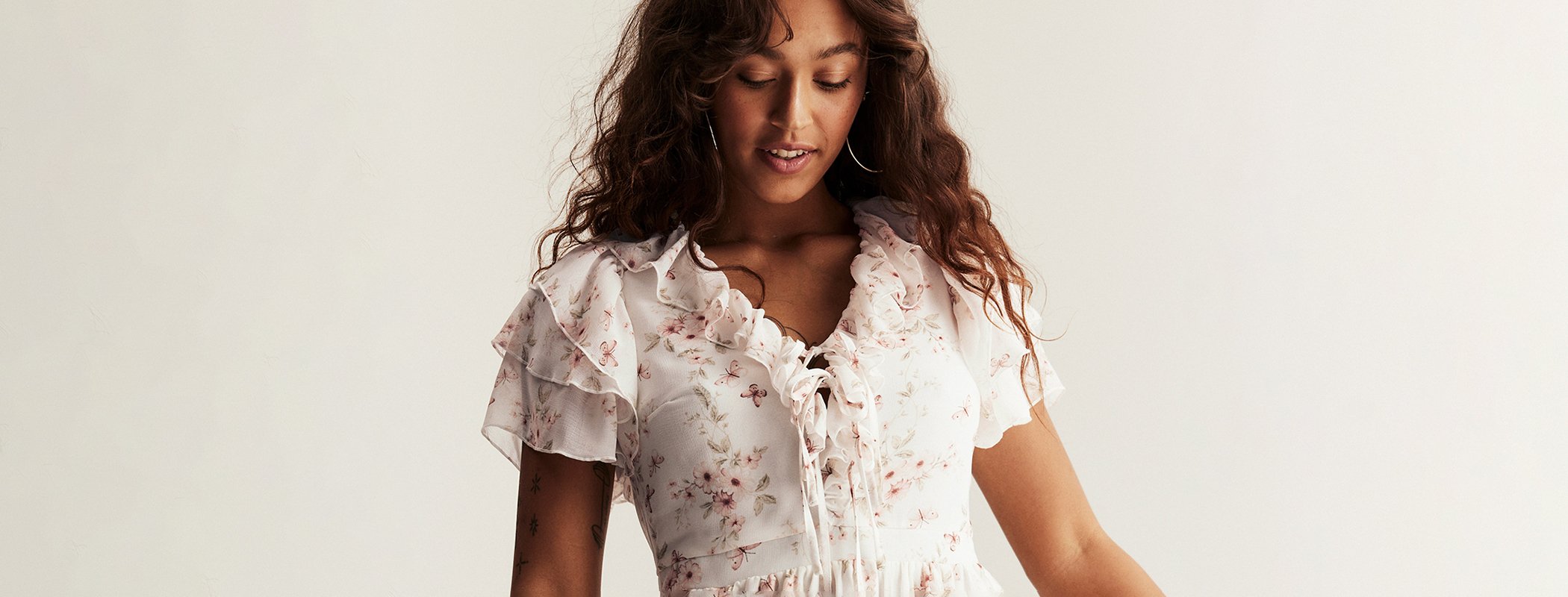 Weißes Baumwollkleid mit Blumenmuster und kurzen Puffärmeln, getragen von einer lächelnden, dunkelhaarigen Frau in sitzender Pose vor hellem Hintergrund.