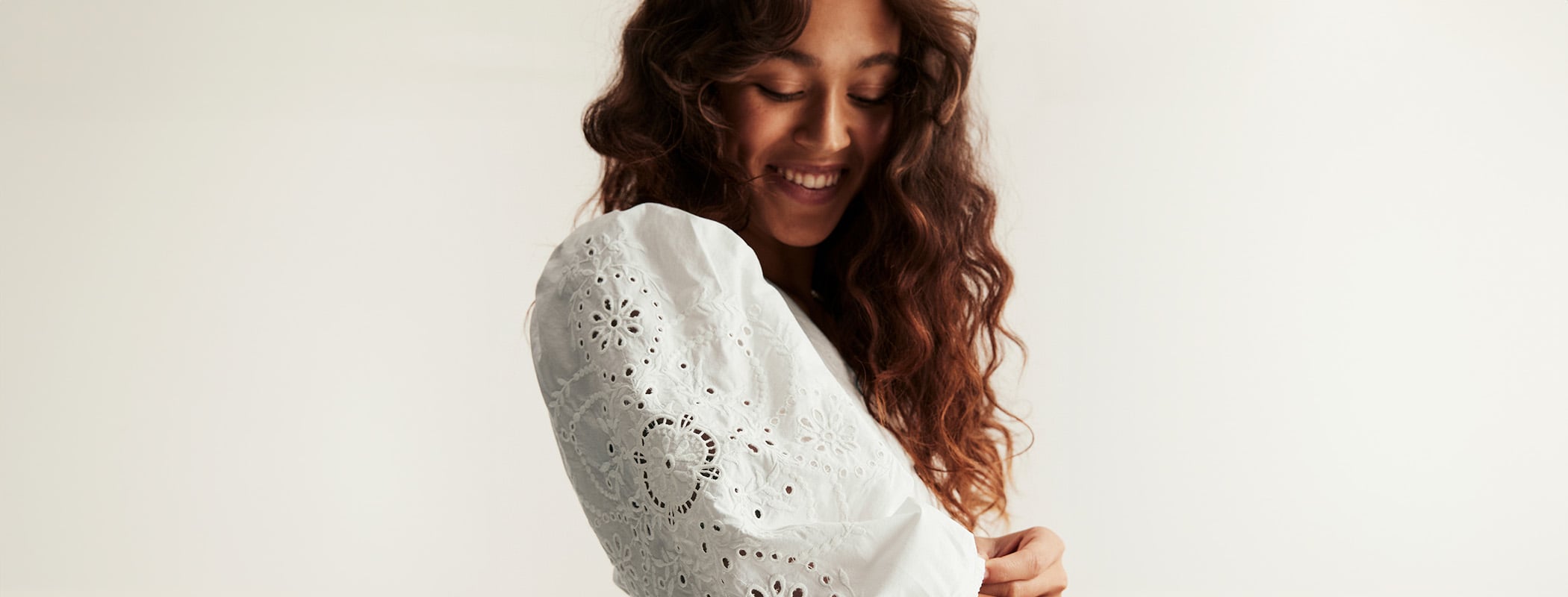 Weiße Bluse mit Lochstickerei-Ärmeln und Puffschultern, Modell richtet den Ärmelbund, langes lockiges Haar, neutraler Hintergrund.