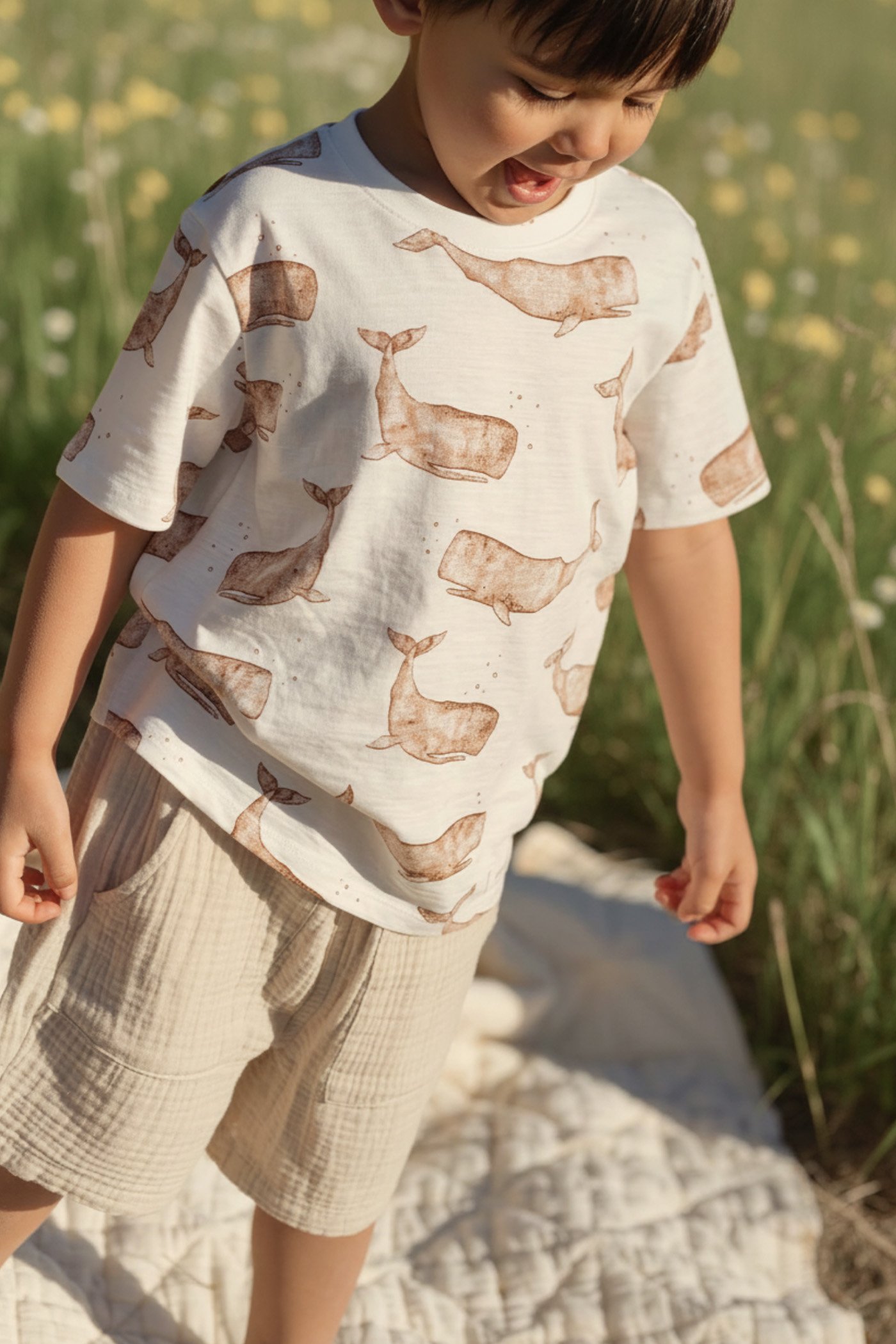 Child wearing a light cotton t-shirt with a soft whale print and beige muslin shorts, photographed outdoors in summer light.