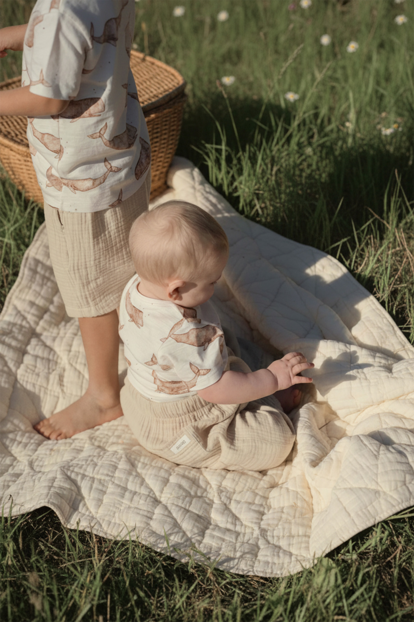 Two children wearing matching cotton outfits with a whale print, one standing and one sitting on a quilt outdoors in a grassy meadow.