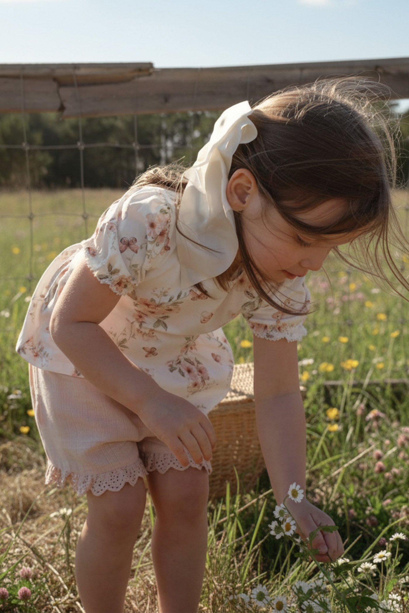 Child wearing a floral cotton outfit with lace details, standing outdoors in a meadow and picking small white flowers.