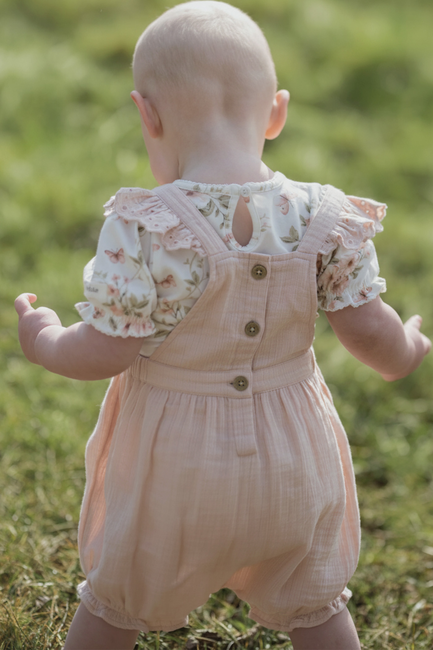 Child wearing light pink cotton dungarees with buttons on the back, layered over a floral short-sleeve top, standing outdoors on grass.