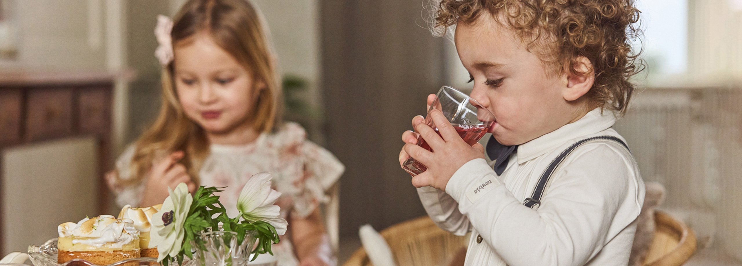 Zwei Kinder an einem gedeckten Tisch mit Kuchen und Blumen, gekleidet in weiche Newbie-Outfits in ruhiger Umgebung.