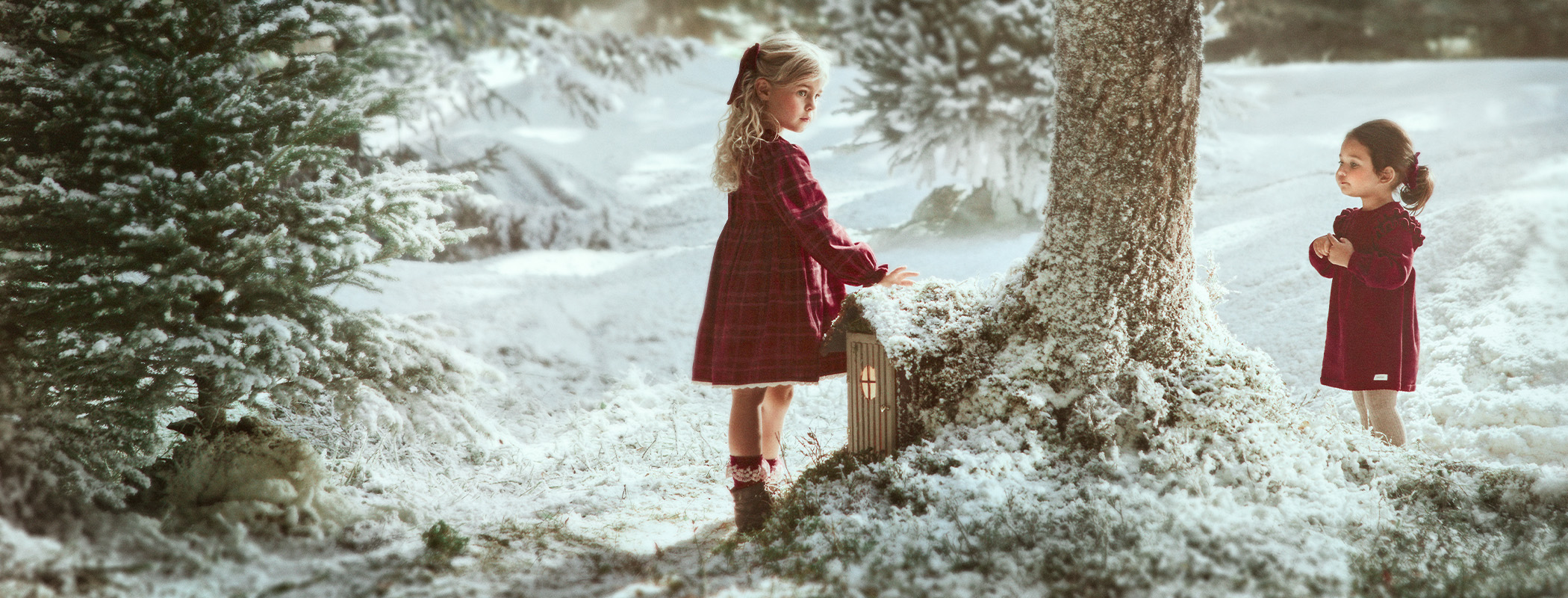 Zwei Kinder in roten Kleidern an einem verschneiten Baum in winterlicher Landschaft