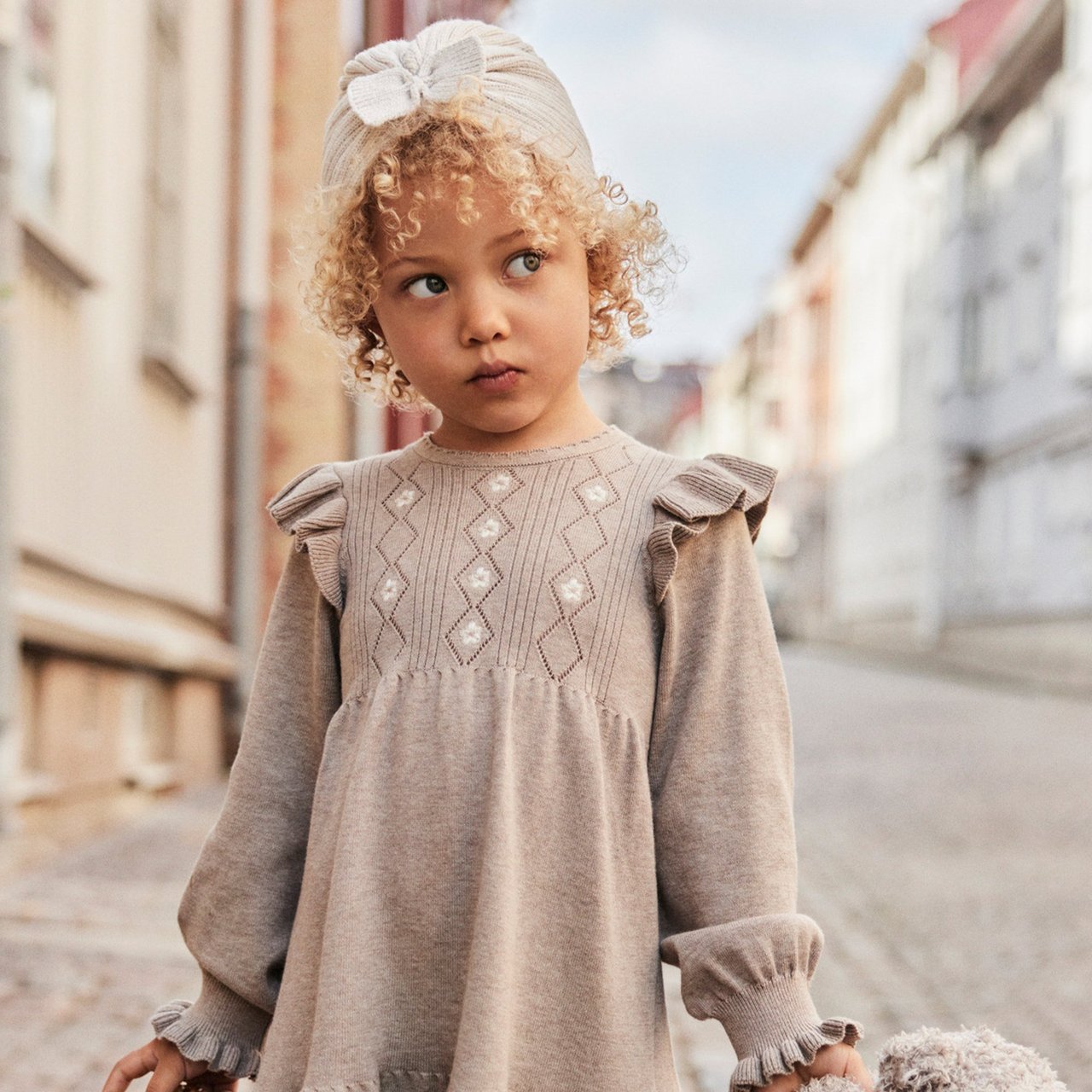 Kind im beigen Rüschenkleid steht auf der Straße und hält etwas Weiches.