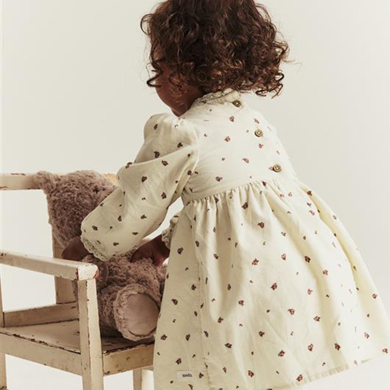Kind im Blumenkleid spielt mit einem Teddybär auf einem Holzstuhl.