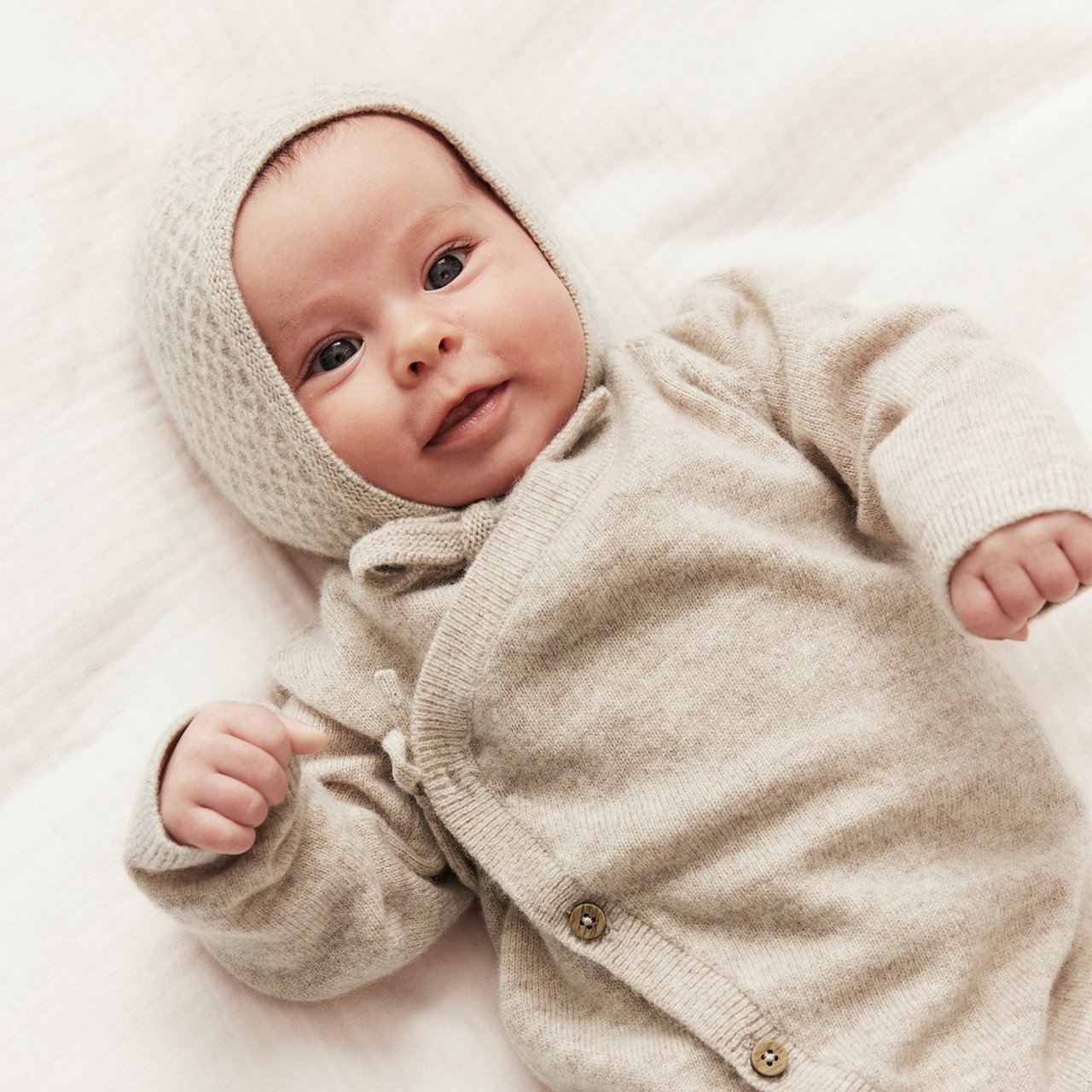 Baby im Strickanzug mit Haube liegt auf einer Decke.