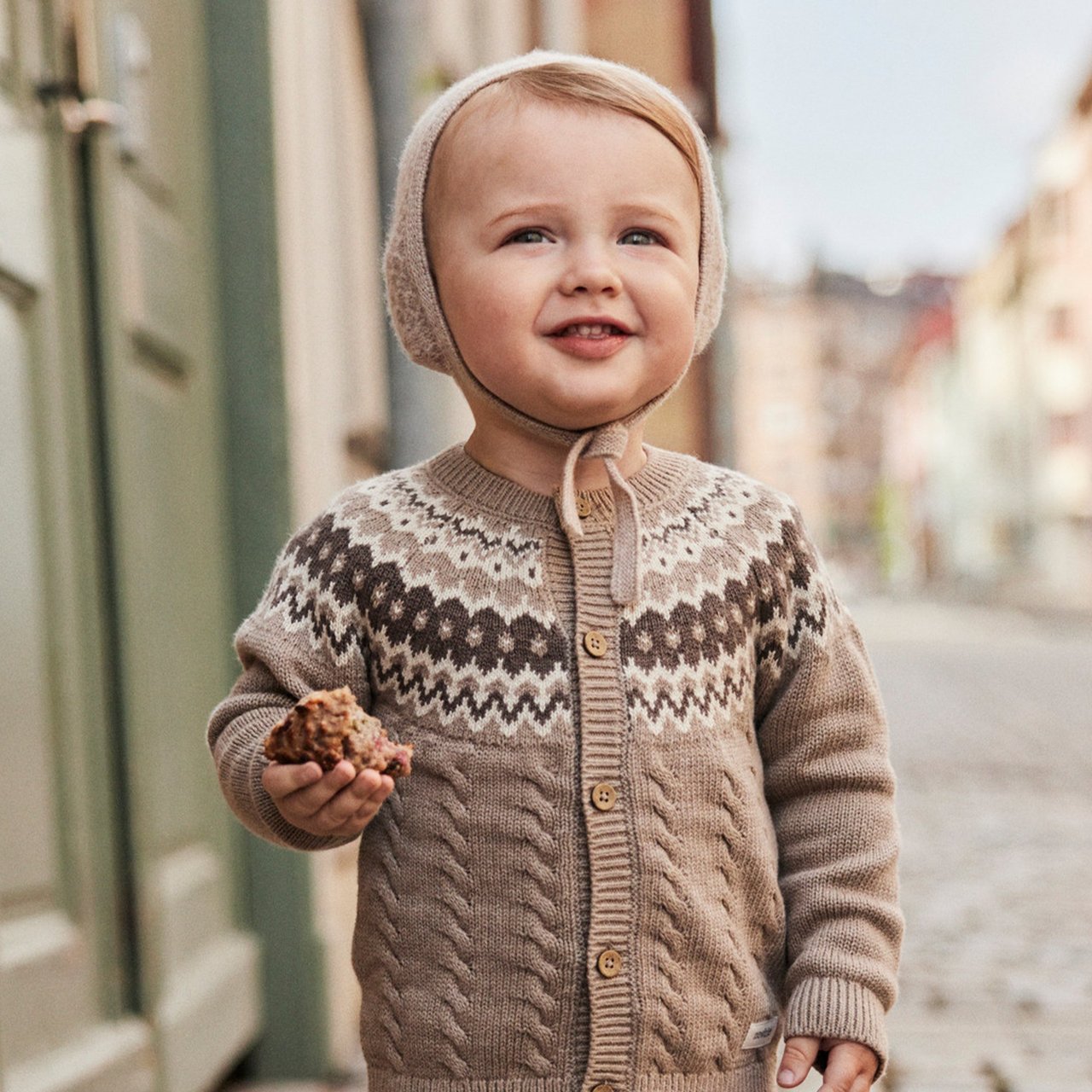 Kleinkind in Strickjacke und Haube geht draußen und hält Essen.
