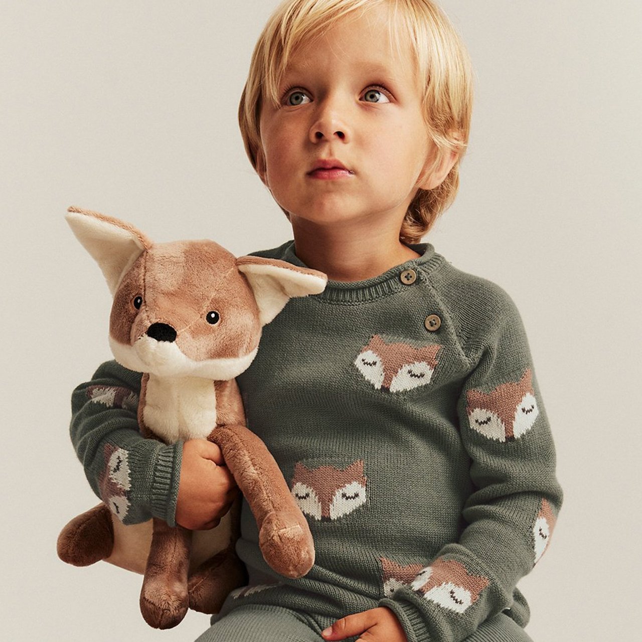 Boy dressed in a green knit embroidered with foxes, holding a toy fox
