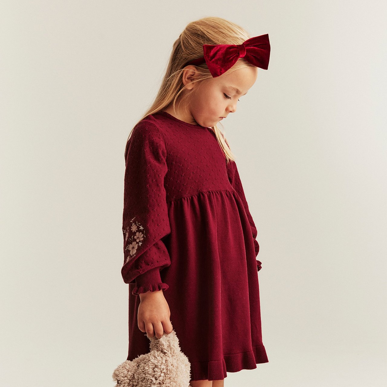 Girl wearing a red christmas dress  with matching red headband rith a bow, holding a teddybear