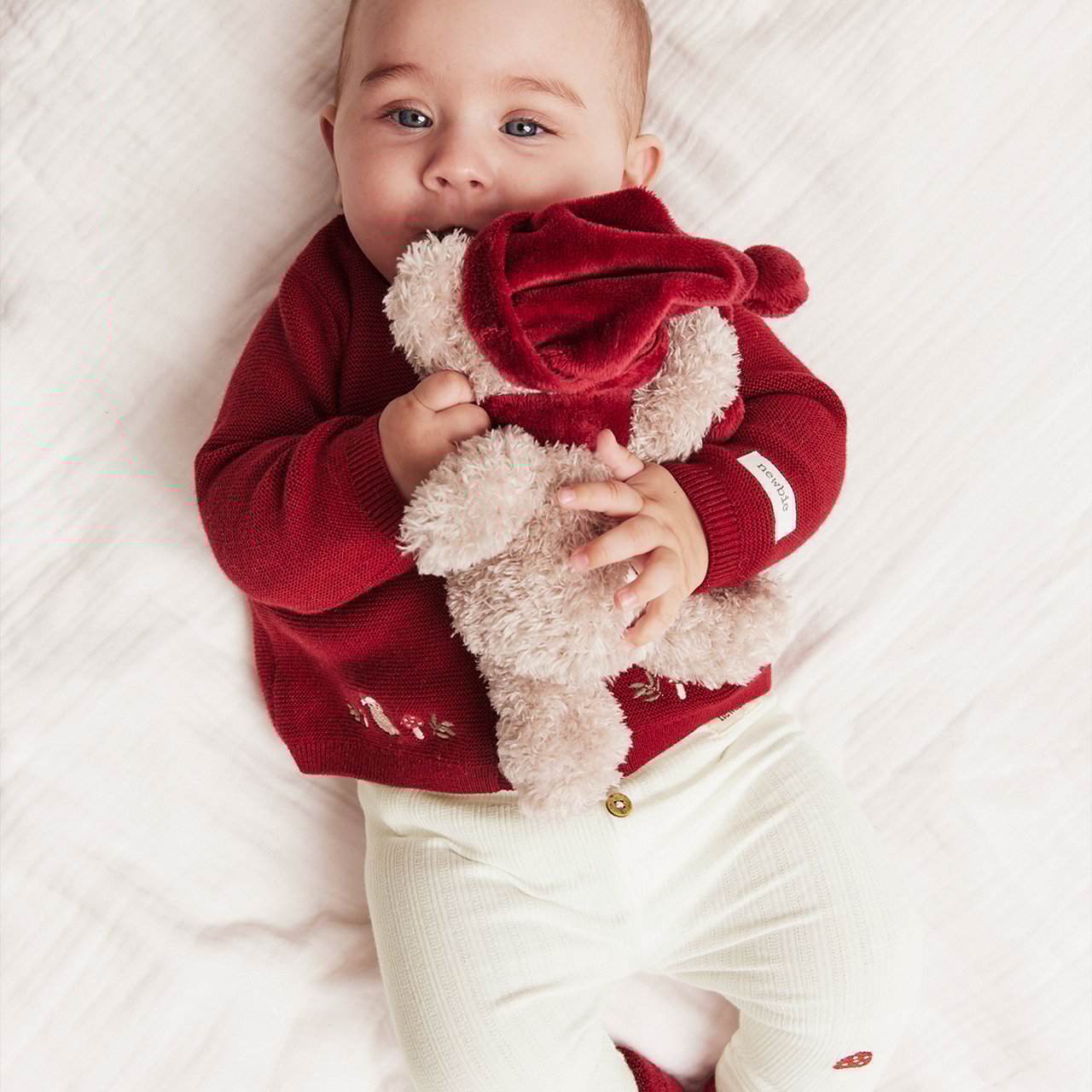 Baby in red knitted christmas sweater holding a teaddybear wearing a santas hat