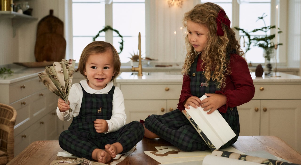 Two children in matching plaid overalls sitting at a kitchen table with Christmas crafts.