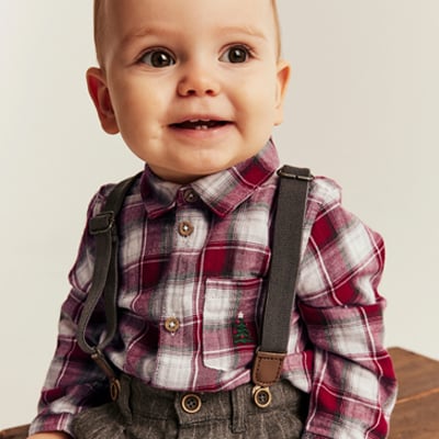 Baby wearing a plaid shirt with suspenders and festive embroidered details.