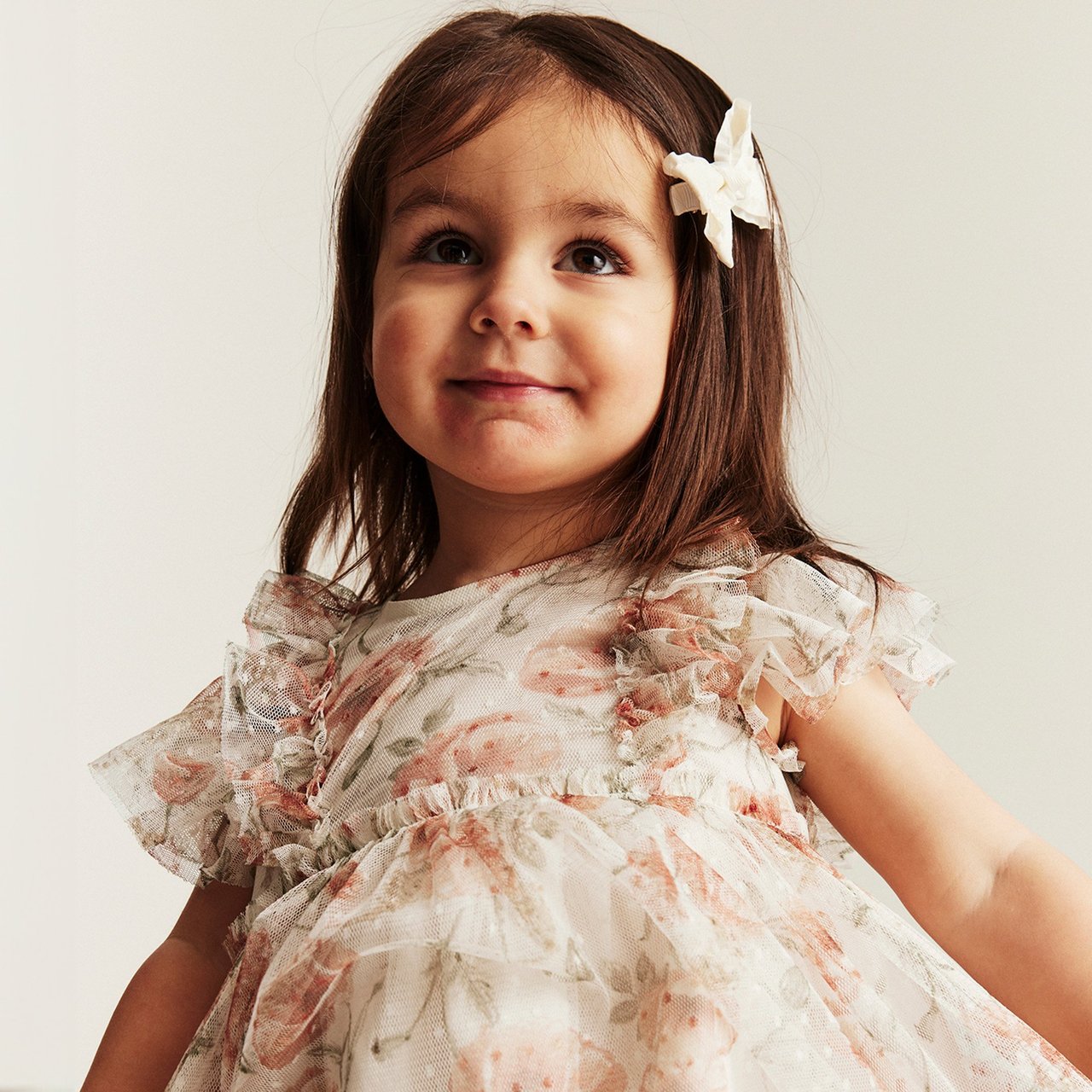 A young child wearing a soft, floral ruffle dress with a light bow in the hair.