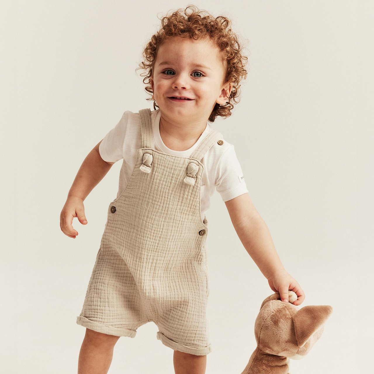 Baby boy wearing beige textured dungaree shorts over a white T‑shirt while holding a soft toy