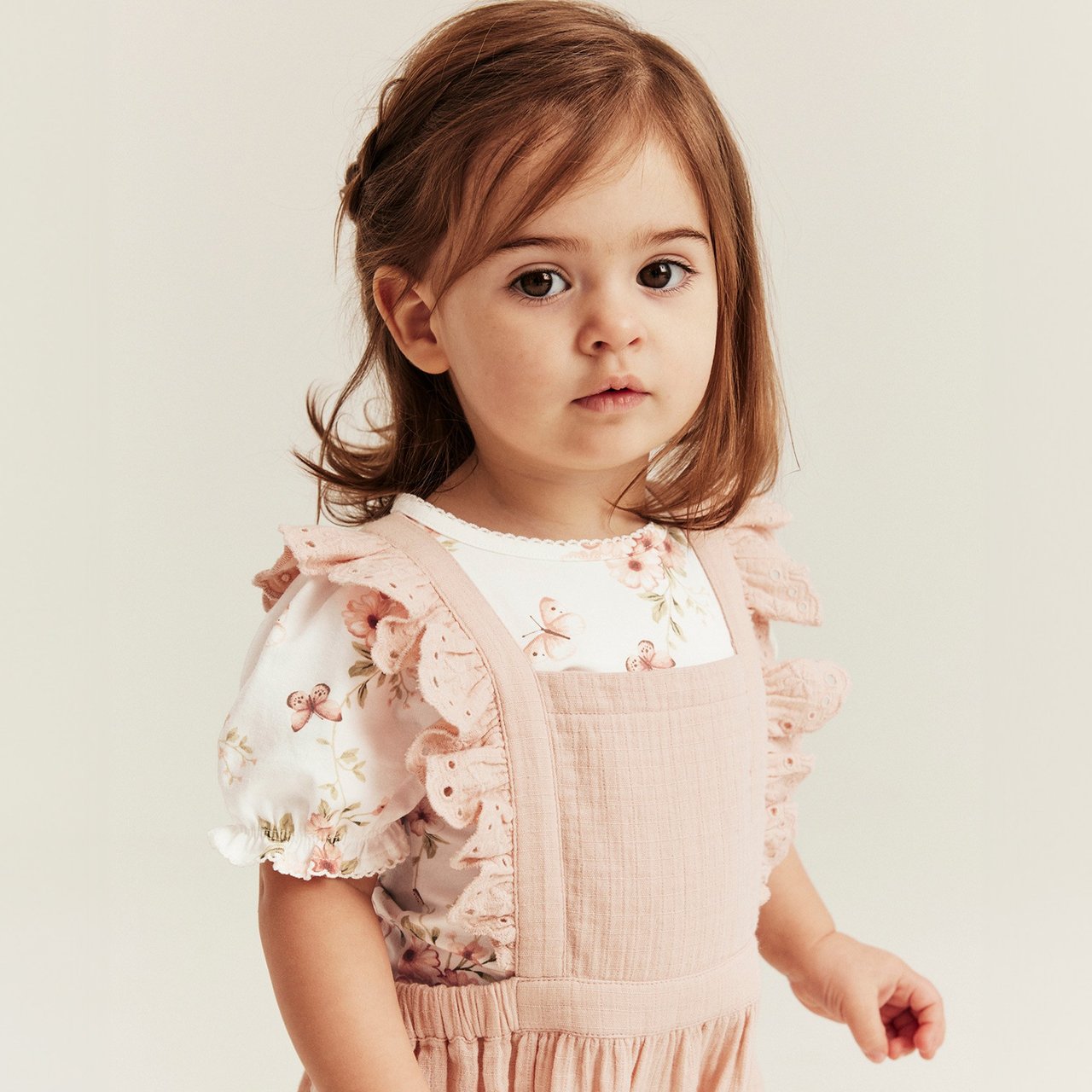Child wearing a light pink pinafore dress with ruffled straps over a white floral top