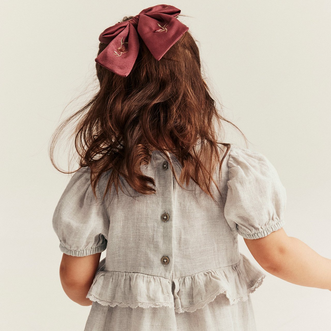 Child seen from behind wearing a light grey linen dress with puff sleeves and a large burgundy hair bow