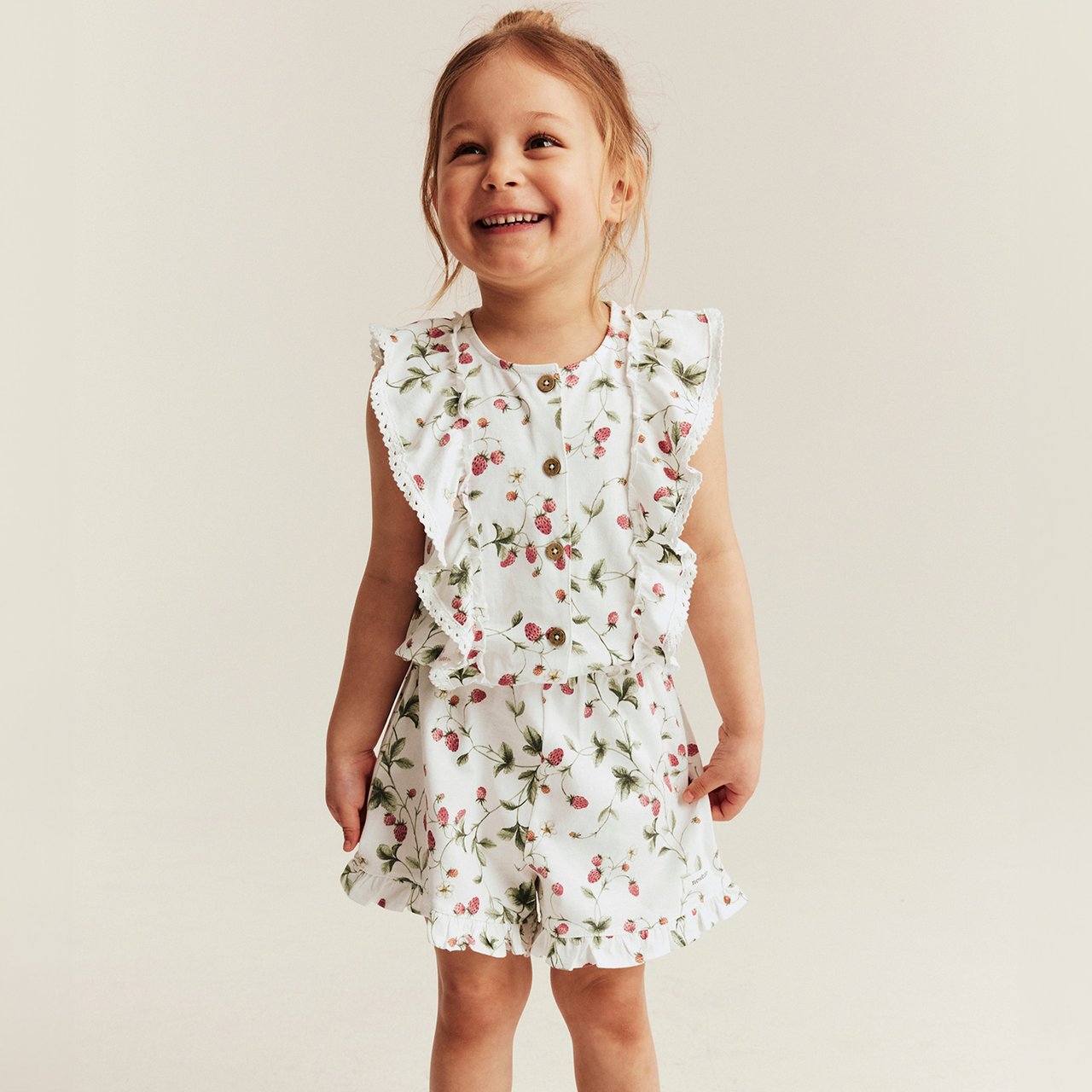 Child wearing a white floral romper with ruffled details and wooden buttons, standing against a light neutral background.