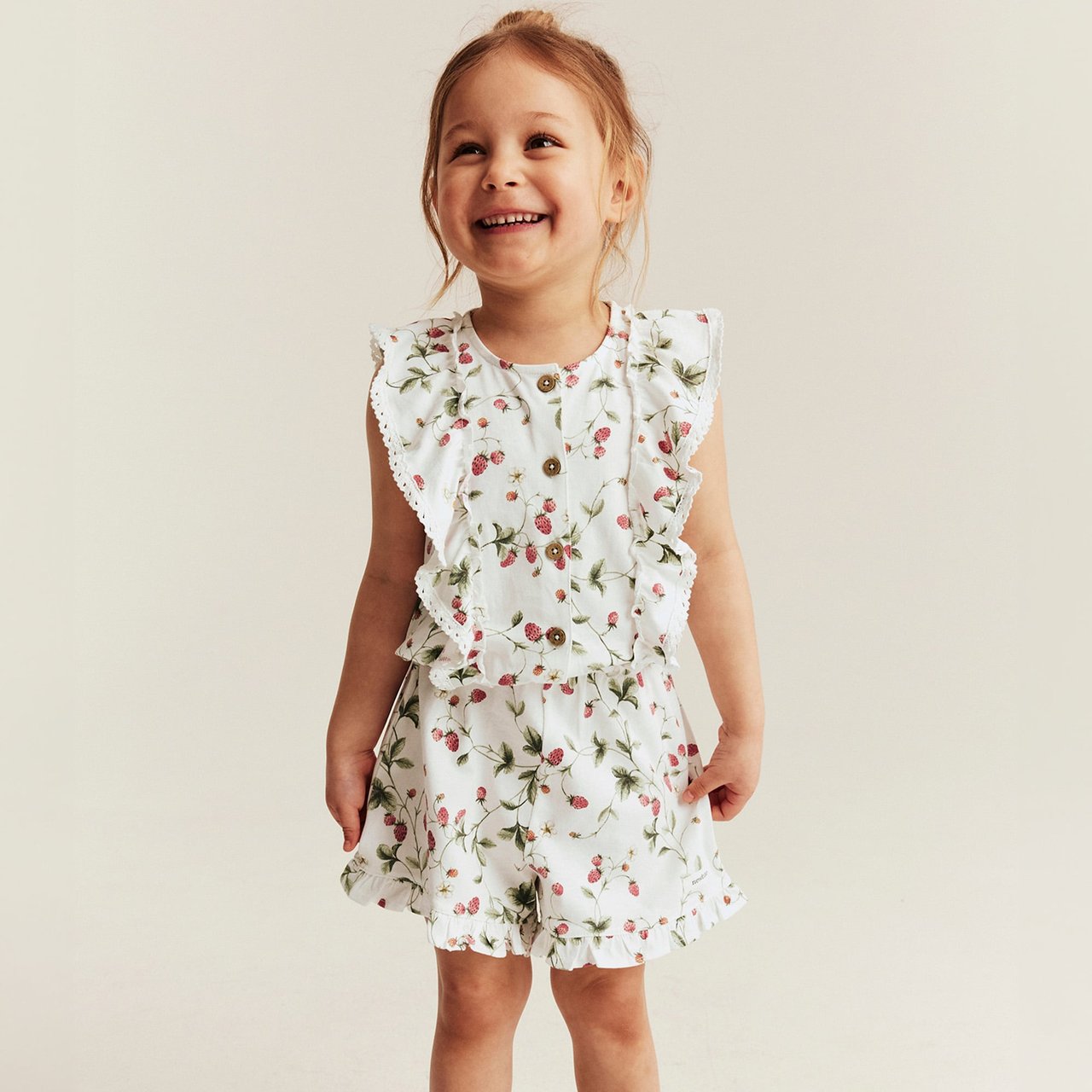 Child wearing a white floral romper with ruffled details and wooden buttons, standing against a light neutral background.
