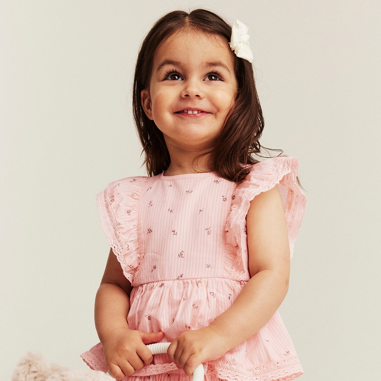 Girl wearing a light pink dress with delicate floral print and ruffled sleeves, standing in soft natural light.
