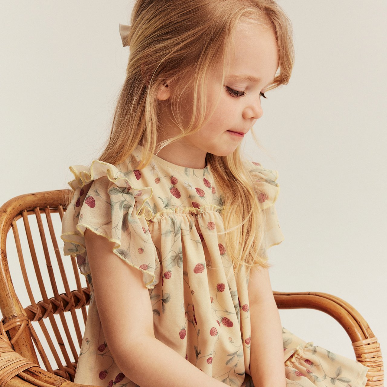 Child wearing a light floral dress with ruffled sleeves, seated on a wicker chair in soft natural light.