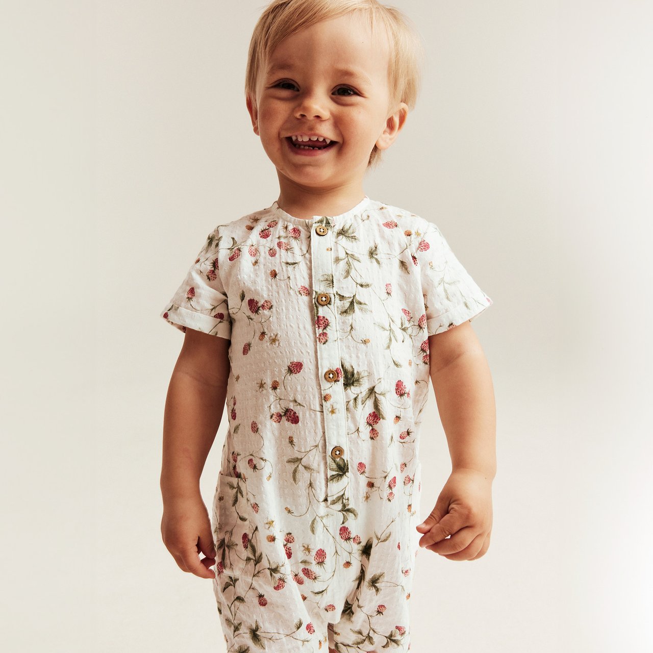 Child wearing a white floral romper with short sleeves and wooden buttons, standing against a light neutral background.