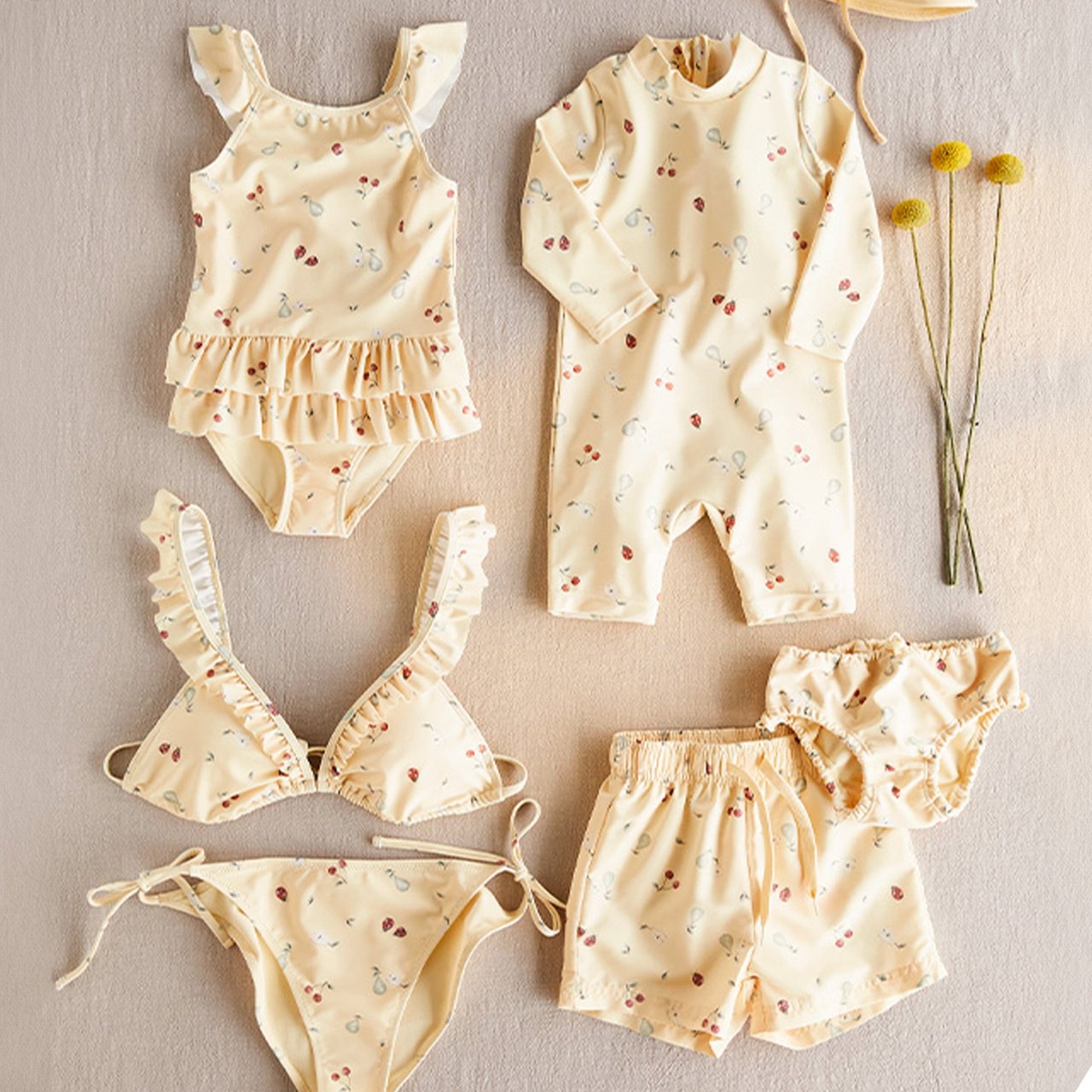 Flat lay of cream-colored baby garments with small floral print, including swimsuits, shorts and a long-sleeve romper on a soft fabric background.