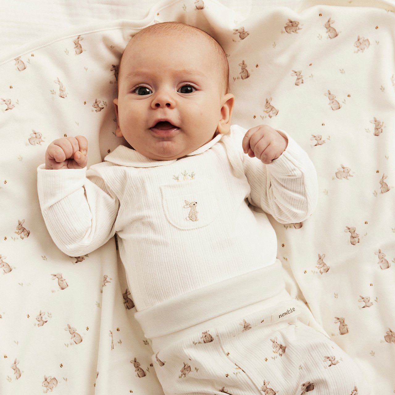 Baby wearing a cream ribbed top and matching pants with small animal print, lying on a soft patterned blanket.