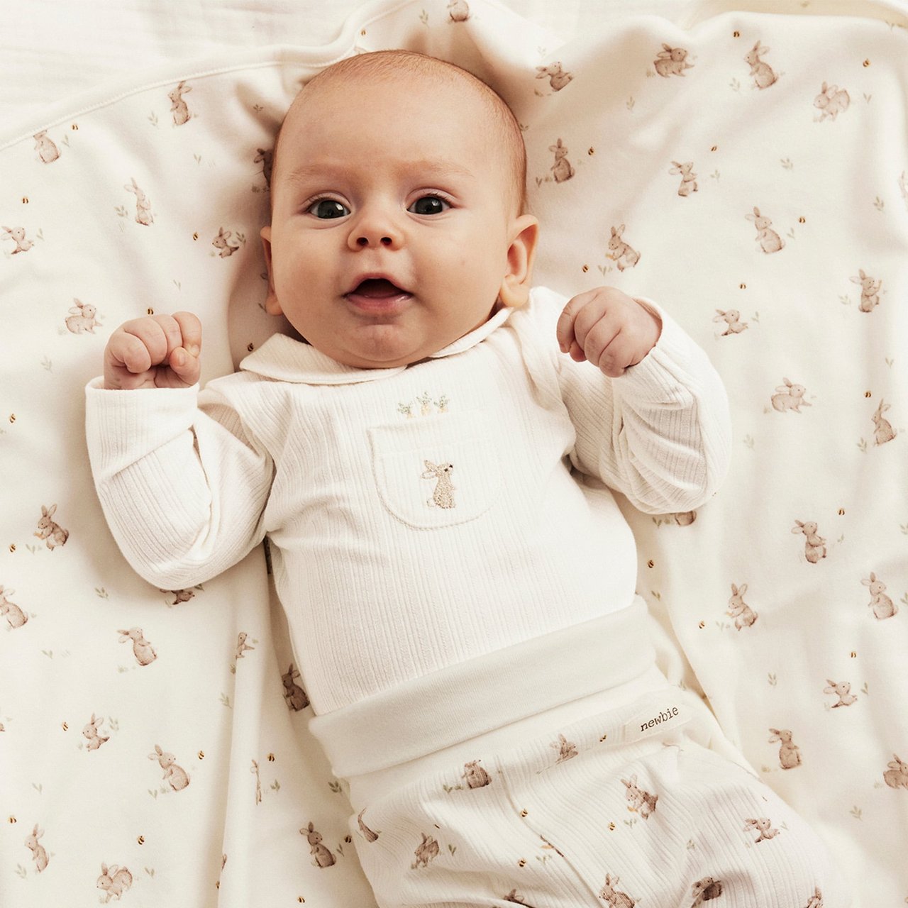 Baby wearing a cream ribbed top and matching pants with small animal print, lying on a soft patterned blanket.
