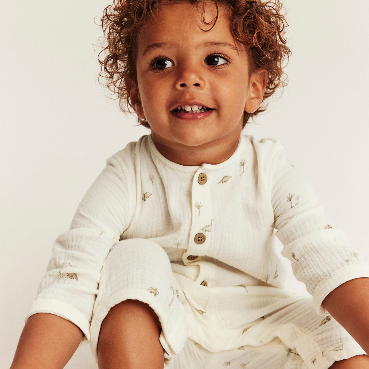 Off-white long-sleeved muslin set with small dinosaur print, front buttons and relaxed fit, worn by child sitting.