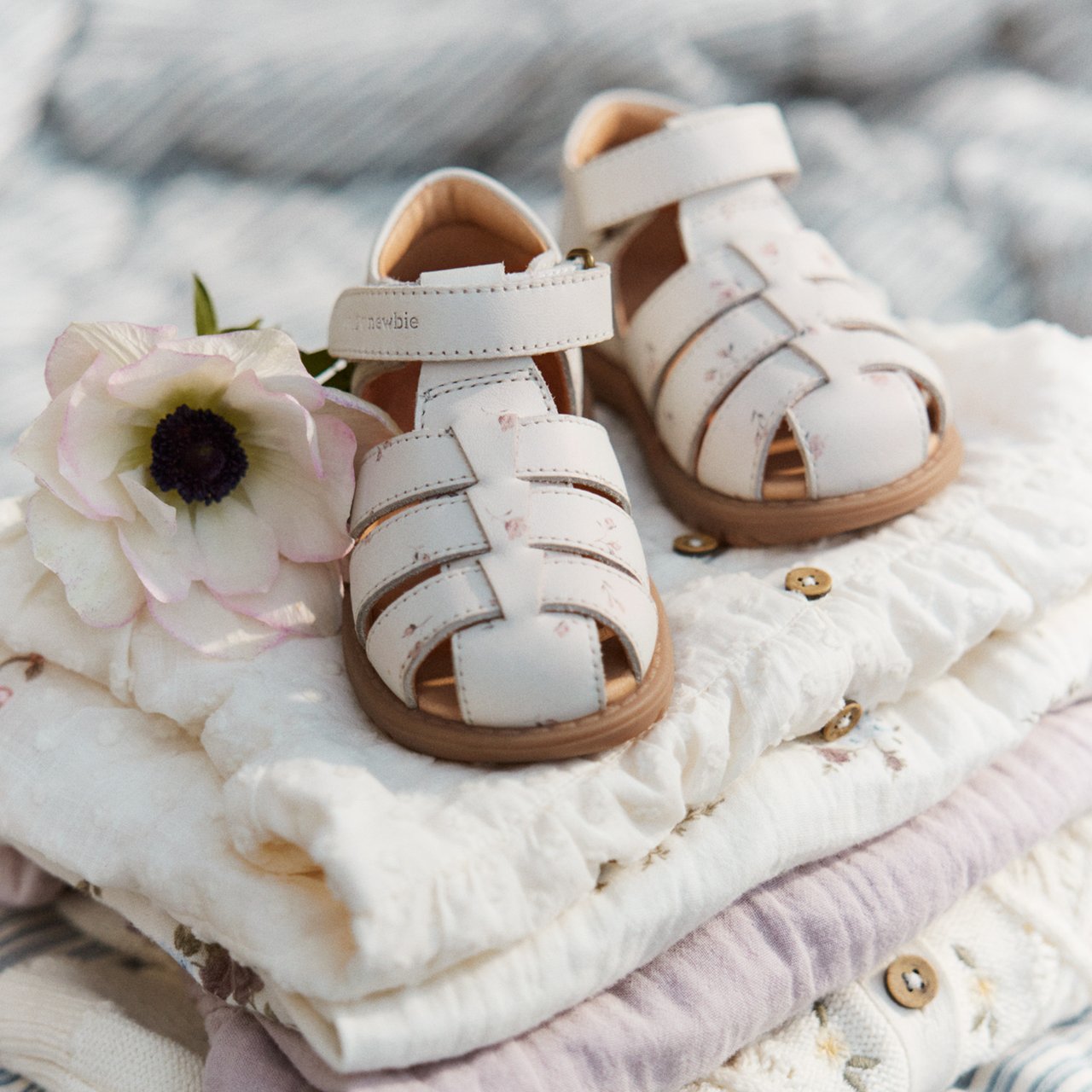 White baby leather sandals with cut-out straps and buckle, placed on folded garments with a flower beside