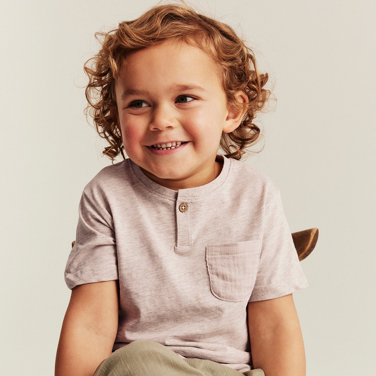Light beige short‑sleeved T-shirt on child, with henley button placket, chest pocket and soft cotton texture.