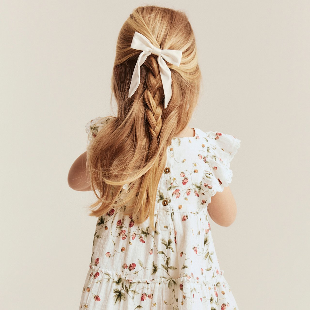 White short-sleeved dress with strawberry print, ruffle sleeves and back buttons, worn by child with braided hair.