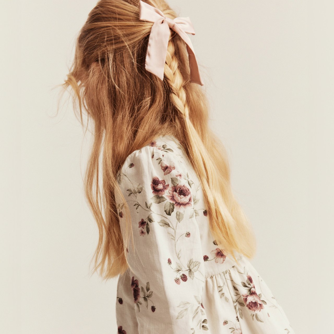 Cream floral dress with long sleeves and braided hair tied with a pink ribbon.