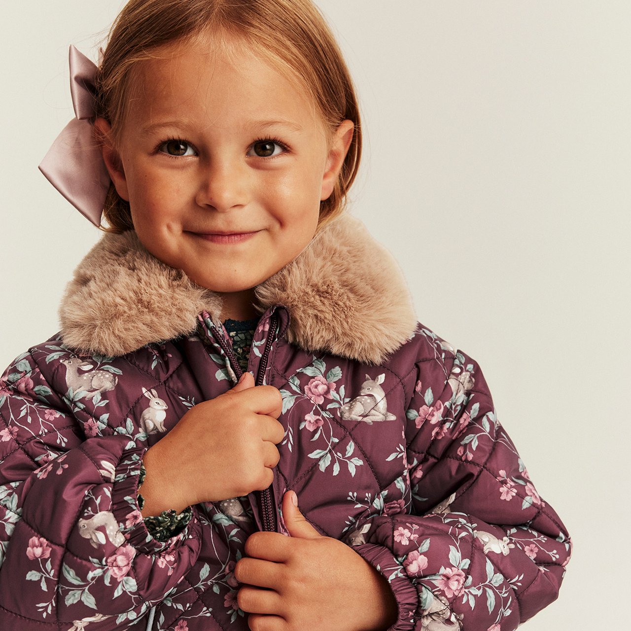 Kind in gesteppter Jacke mit Blumenmuster, Kunstfellkragen und großer Schleife im Haar