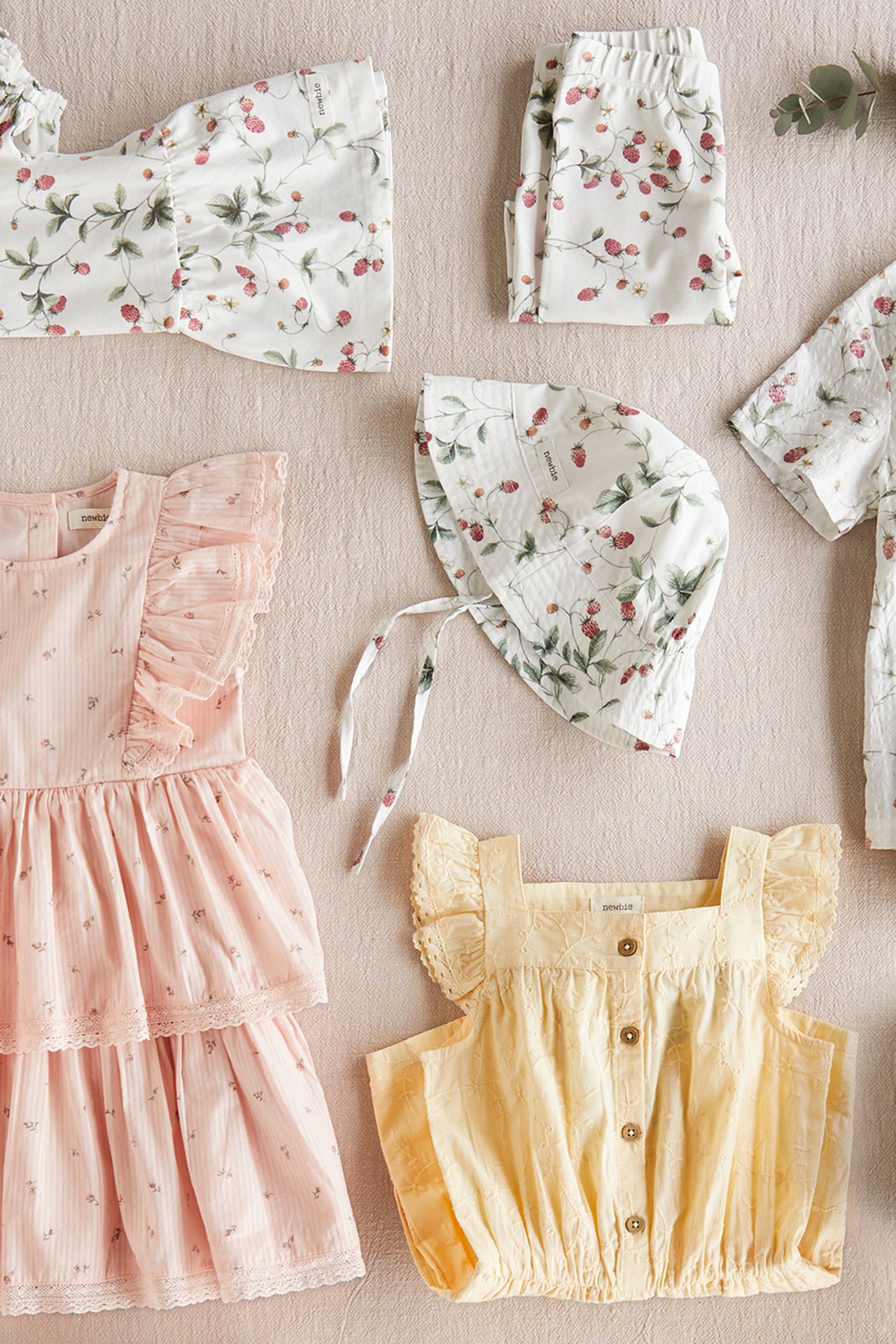 Flatlay mit floraler Babykleidung, rosafarbenem Kleid, gelbem Oberteil und passender Haube auf neutralem Hintergrund
