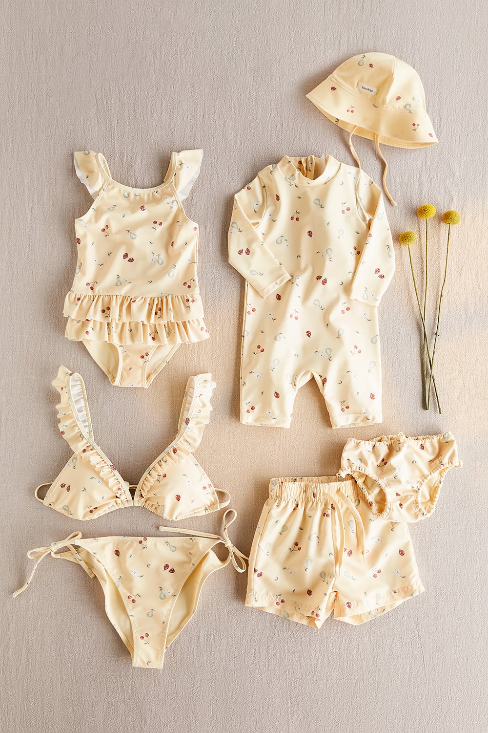 Flat lay of baby swimwear in soft cream with floral print, including swimsuit, bikini, swim shorts and sun hat.