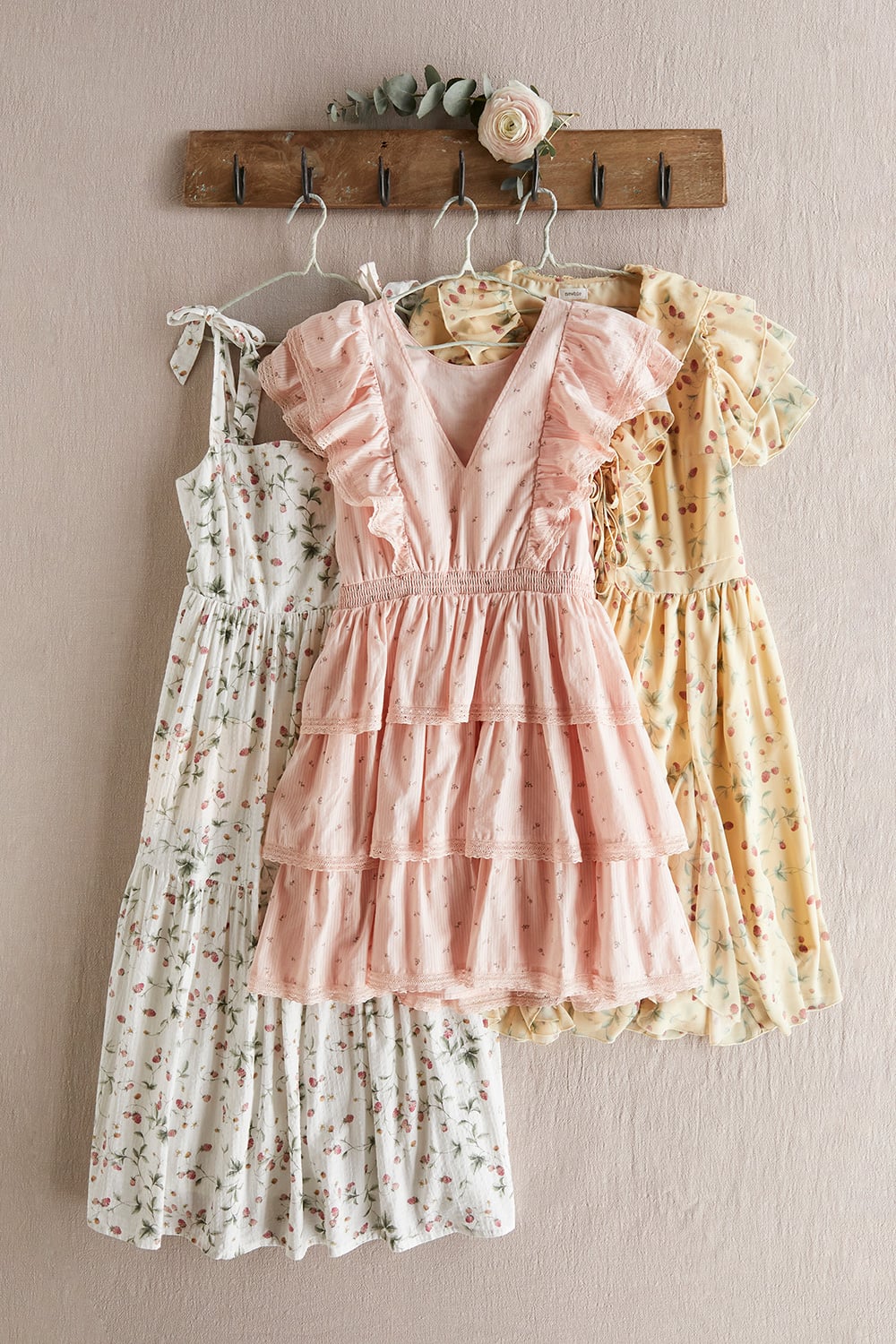 Three women’s dresses in soft floral prints hanging on a wooden rack in muted tones of cream, blush and yellow.