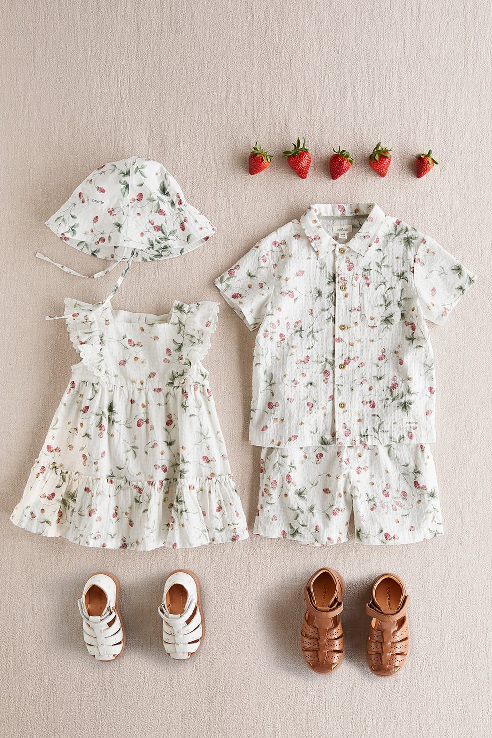 Flat lay of baby outfit in soft cream with floral print, dress, short-sleeve shirt, shorts, sun hat and leather sandals.