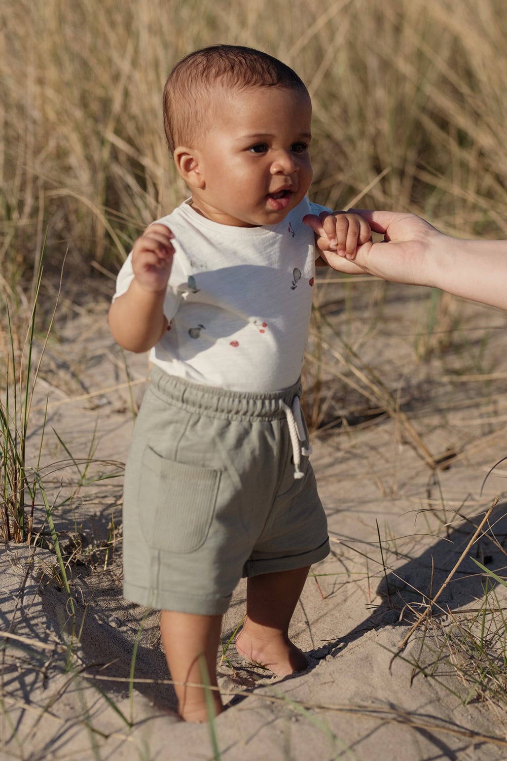 White short‑sleeved bodysuit with small fruit print and green shorts with drawstring, worn by child on sandy beach.