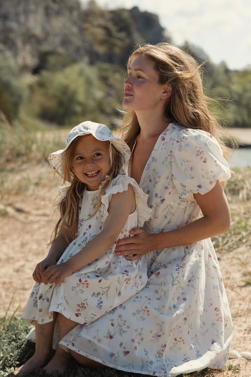 Two people in light floral dresses, sitting together on sandy beach with natural landscape background.