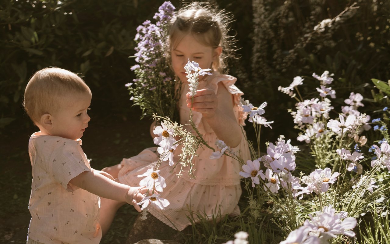 Zwei Kinder in zarten rosafarbenen Sommeroutfits zwischen Wildblumen im Sonnenlicht.