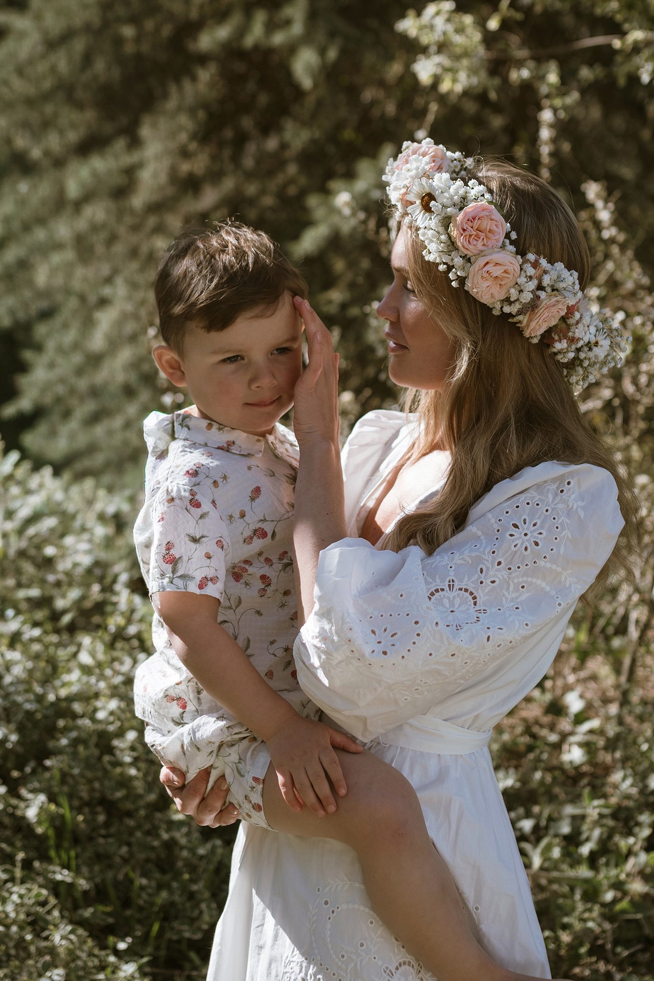 Eine erwachsene Person im weißen Kleid mit Blumenkranz hält ein Kind im sommerlichen Grün.
