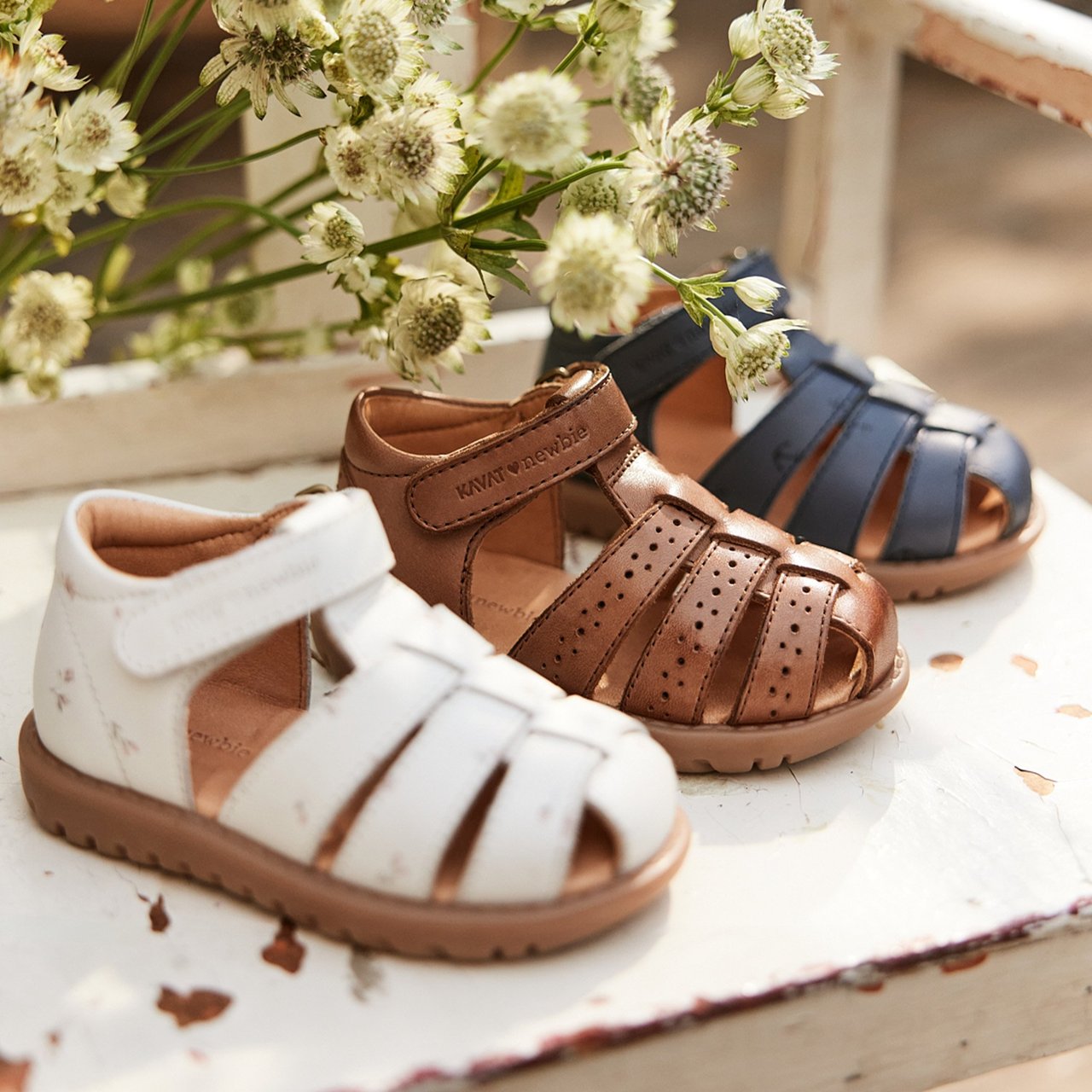Children’s leather sandals in white, brown and navy, arranged on a vintage white bench with soft flowers in the background.