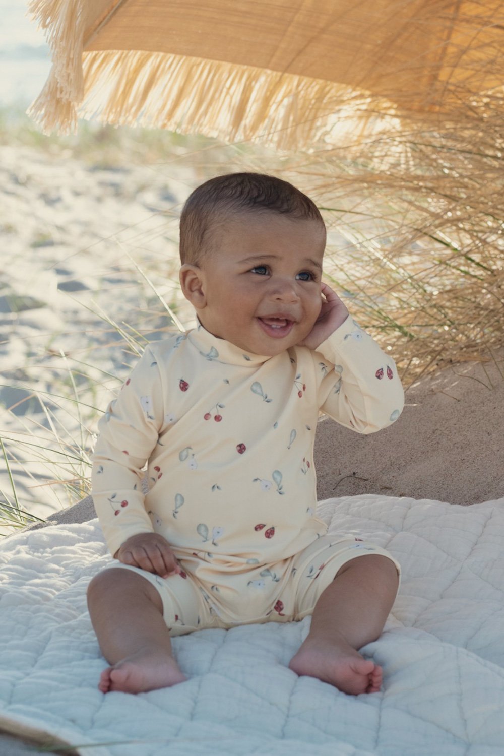 Baby sitting on a quilted blanket wearing a cream long-sleeve top and shorts with a small fruit print.