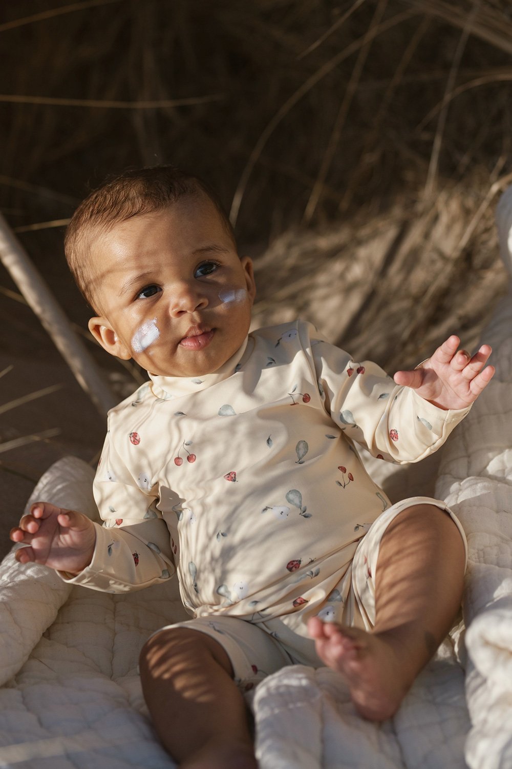 Baby sitting on a quilted blanket wearing a butter yellow long-sleeve UV-suit with an all-over fruit print.