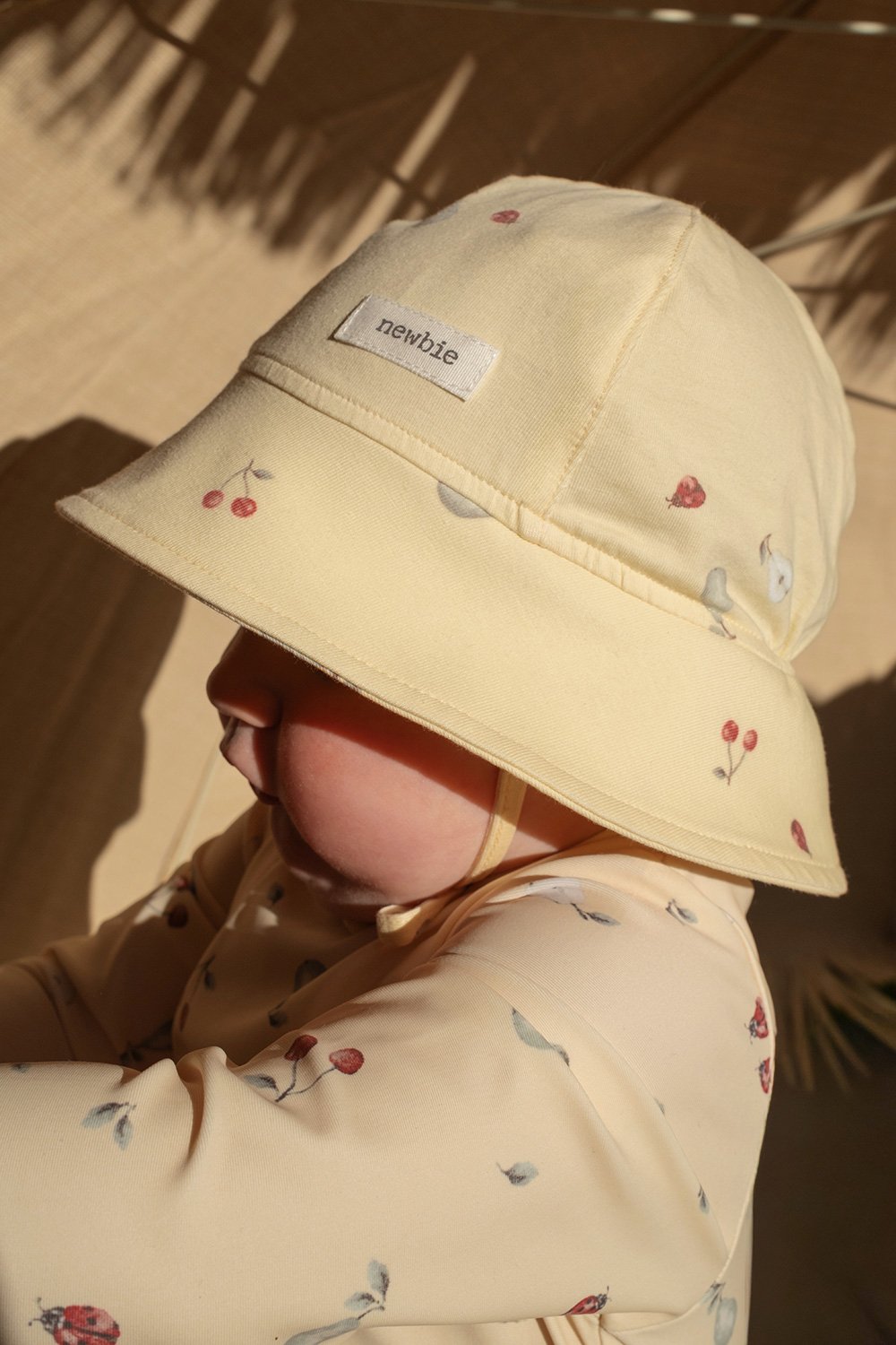 Baby wearing a yellow bucket hat with fruit print and a visible Newbie label on the front.