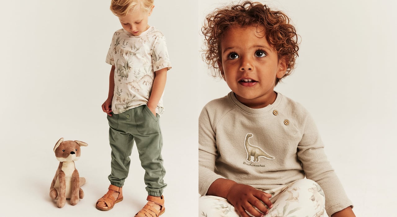Toddler in beige long sleeve top with dinosaur print and matching pants, alongside another child in printed T-shirt and green trousers with soft toy.
